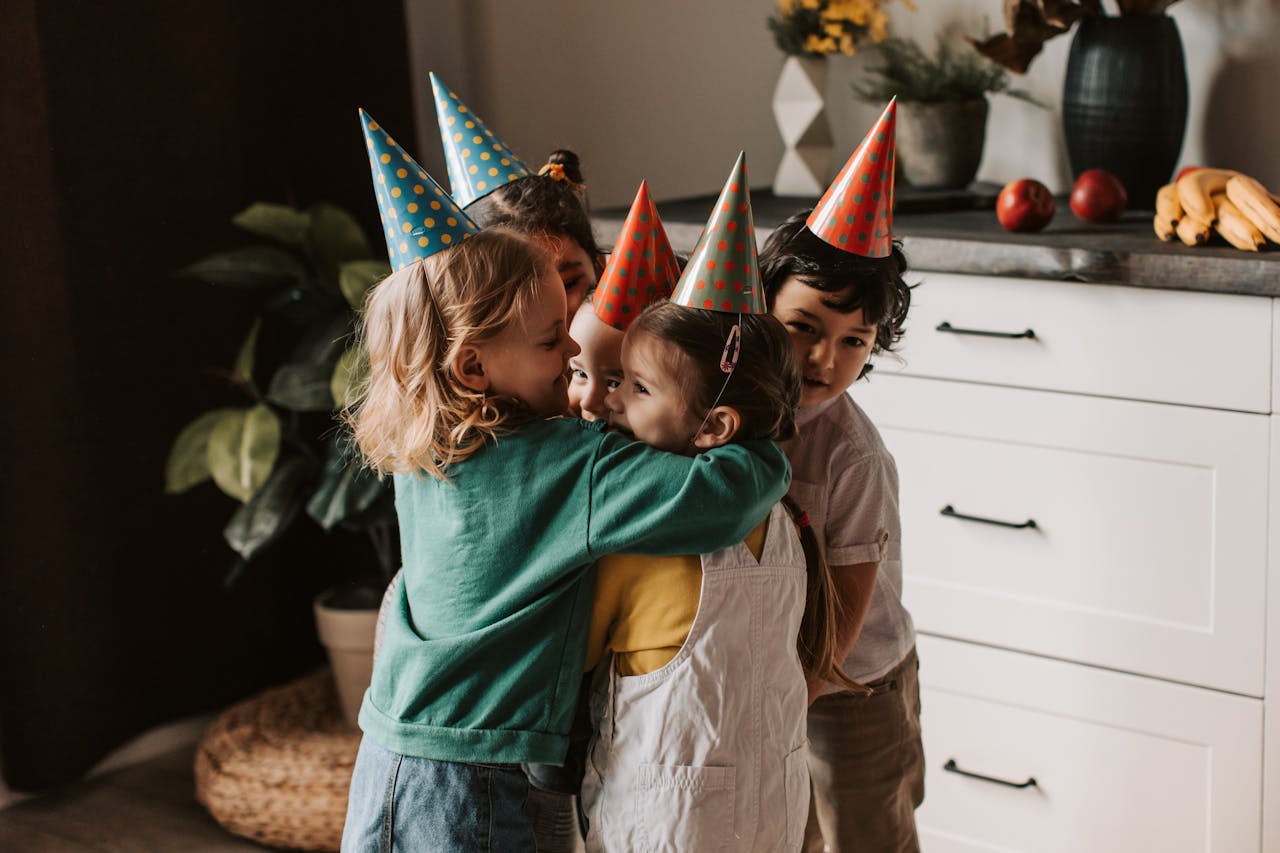 gallery-03 Four kids wearing party hats and hugging warmly during a birthday celebration indoors.