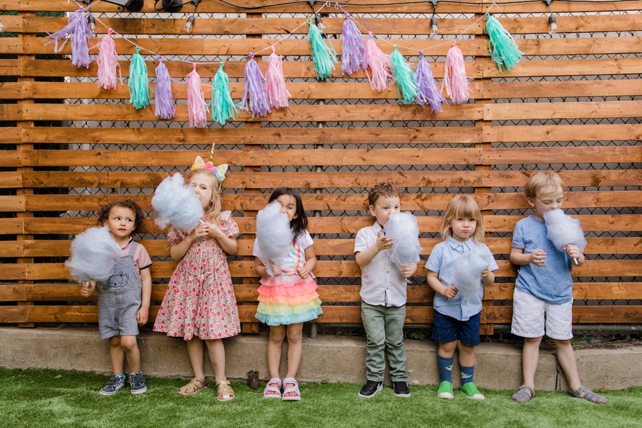 gallery-04 Kids enjoying cotton candy at an outdoor celebration with pastel decorations against a wooden fence.